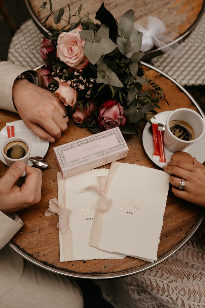 Couple’s hands around coffee, roses and ribboned stationery on a café table in warm light