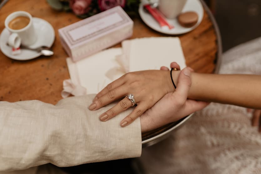 Close-up of a couple holding hands at a wooden table, with a wedding ring and soft warm light