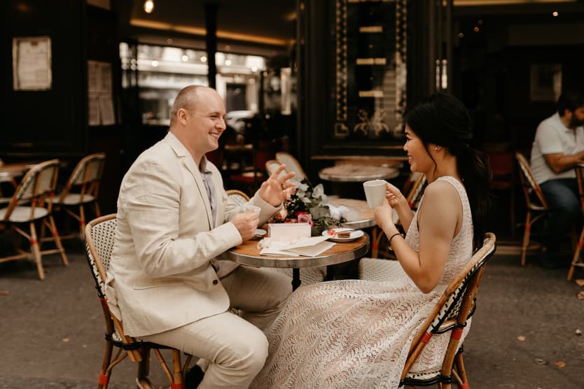 Couple in wedding attire shares coffee at a Paris café, warm light on the small table and flowers