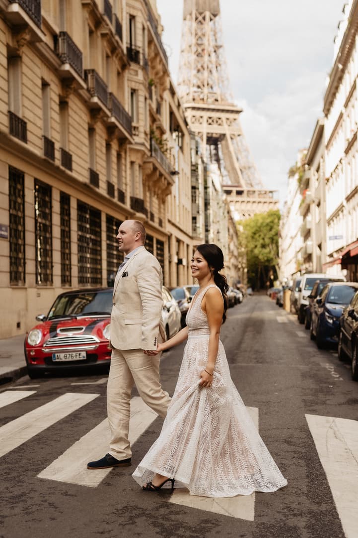 Bride and groom cross a Paris street with the Eiffel Tower rising behind them in soft light