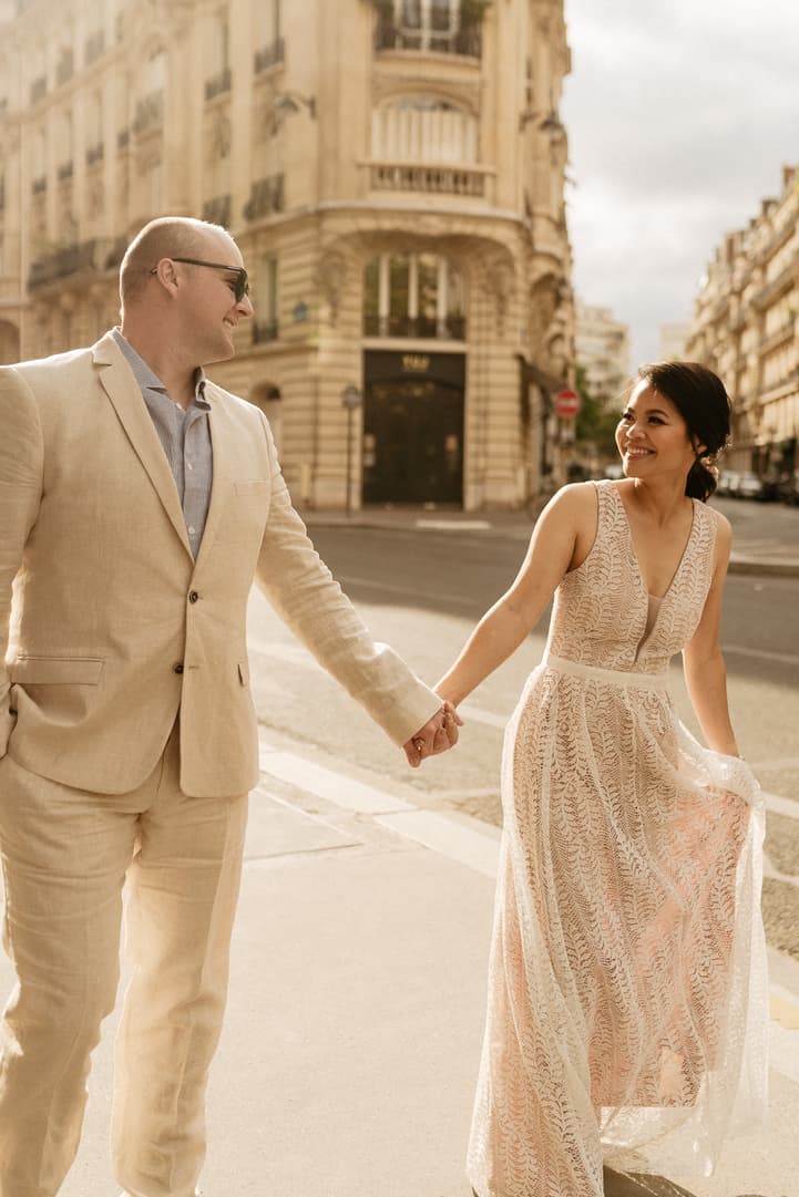 A bride and groom hold hands on a Paris street in warm evening light beside ornate buildings