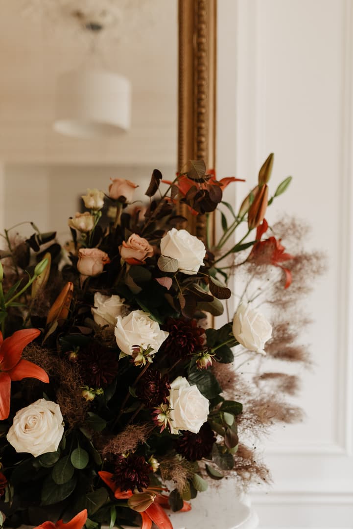 Soft daylight illuminates a lush bridal bouquet of white roses and rust lilies beside a gilt mirror