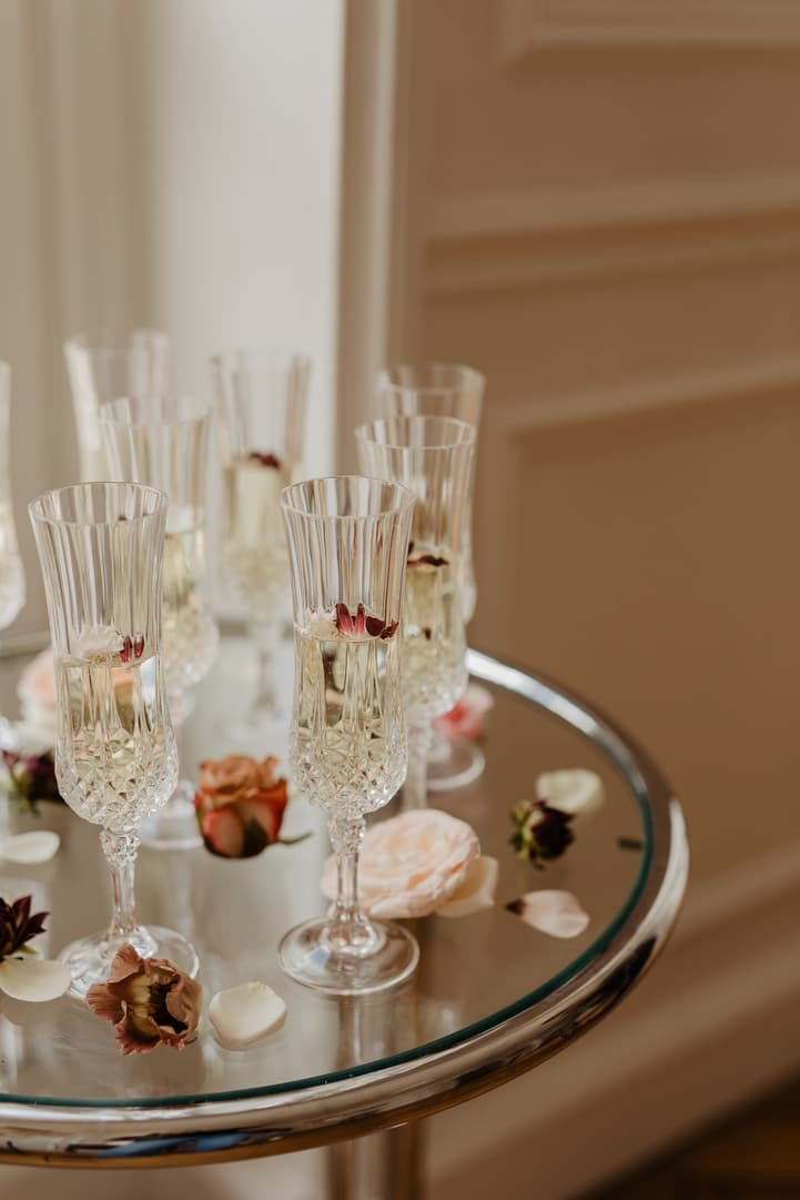 Champagne flutes and scattered rose petals rest on a mirrored tray in soft window light