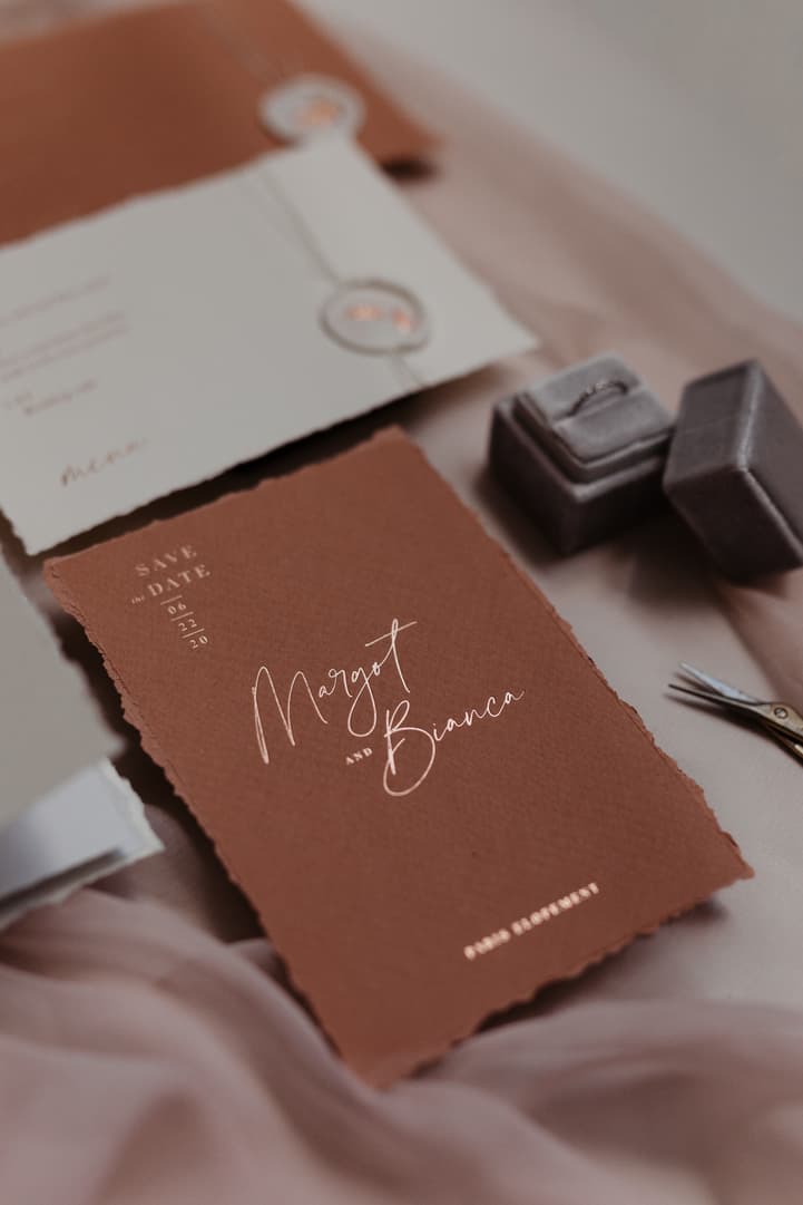 Elegant wedding stationery and ring boxes on soft fabric, with a brown save-the-date card centerpiece