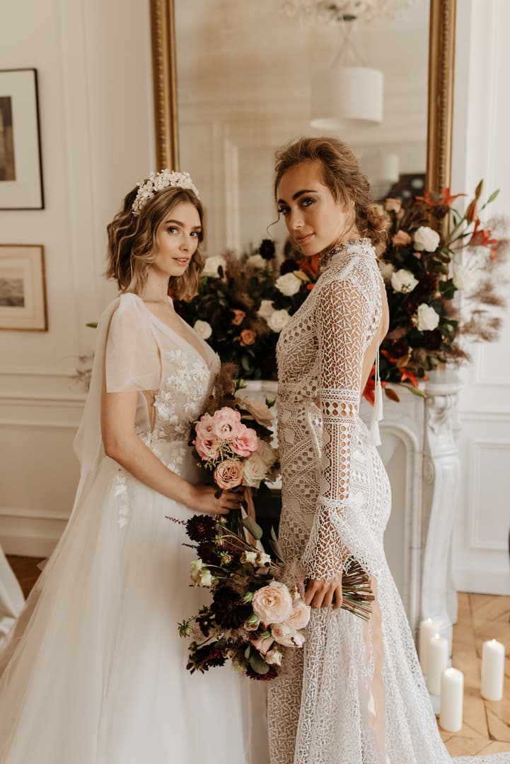 Two brides pose in a Paris room, holding bouquets beside a flowered mantel in soft window light