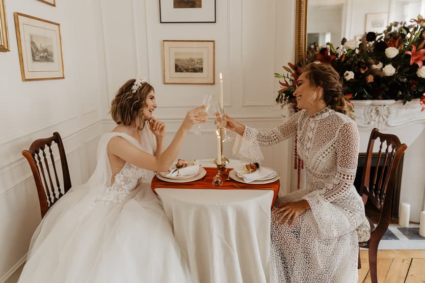 Bride in white gown and lace-clad partner toast at a small table in a sunlit Paris room beside flowers