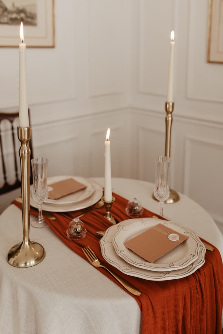 Candlelit Paris table setting with gold holders, blush plates, and a rust runner in a bright room