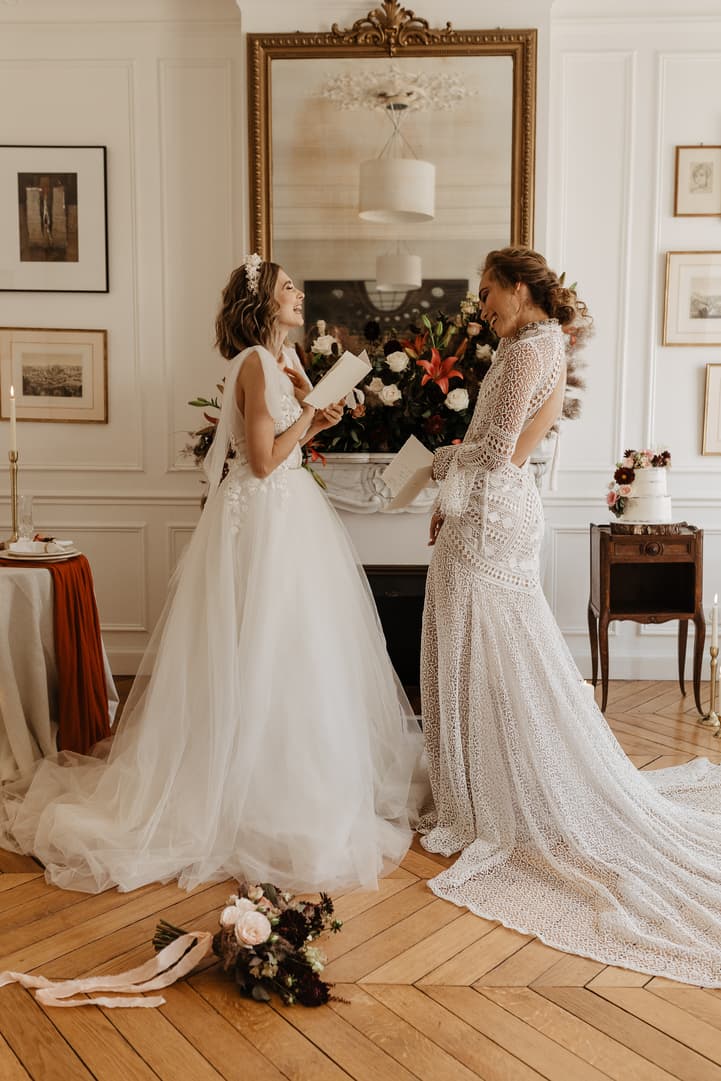 Bride in a white gown speaks with a woman in lace by a floral mantel in a Paris hotel room