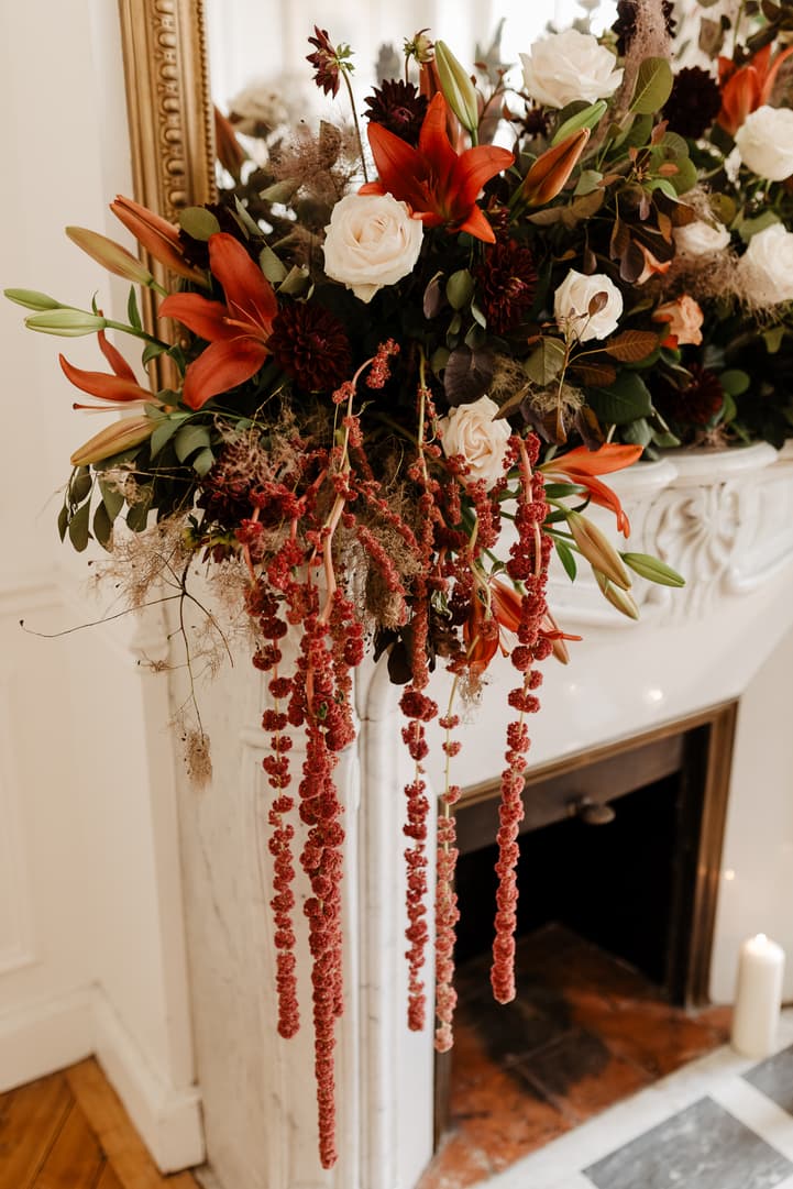 Autumnal wedding flowers cascade over a white marble fireplace in warm indoor light