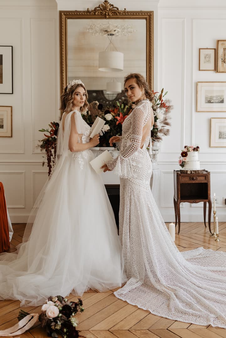 Two brides in lace gowns stand in soft daylight beside a mantel, candles and a cake in a Paris room
