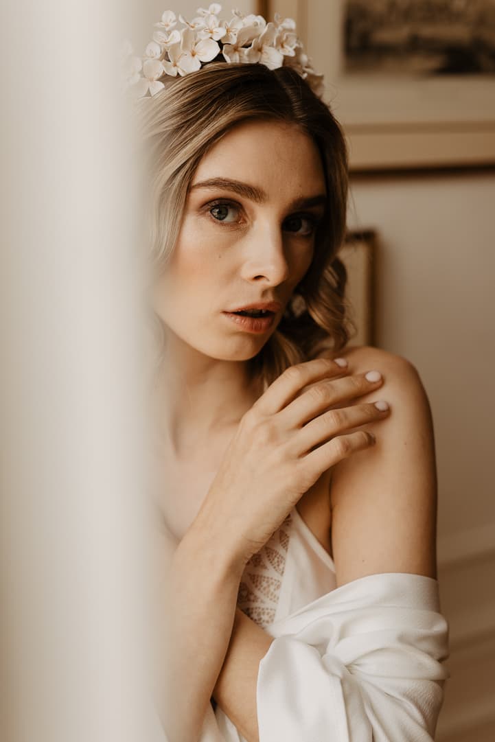 Bride with flower crown adjusts her shoulder in soft window light inside a hotel room