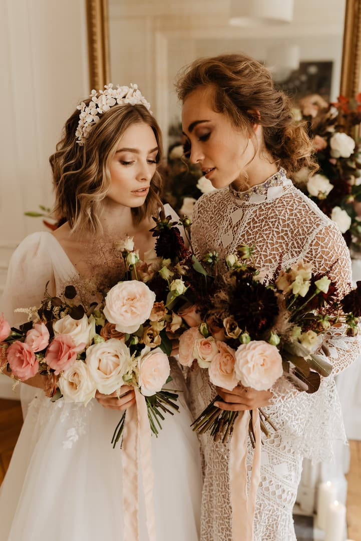 Two brides in lace and tulle hold pastel bouquets in soft indoor light before a mirror