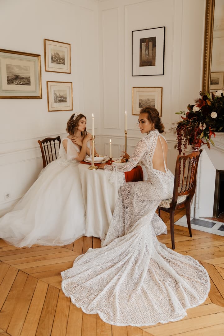 Two women in lace wedding dresses sit at a candlelit table in a Paris hotel room