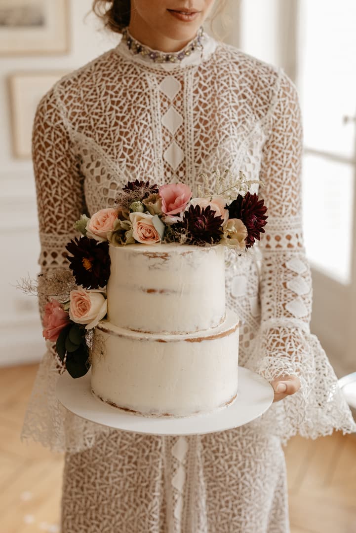 Bride in lace dress holds a two-tier cake topped with roses in soft window light