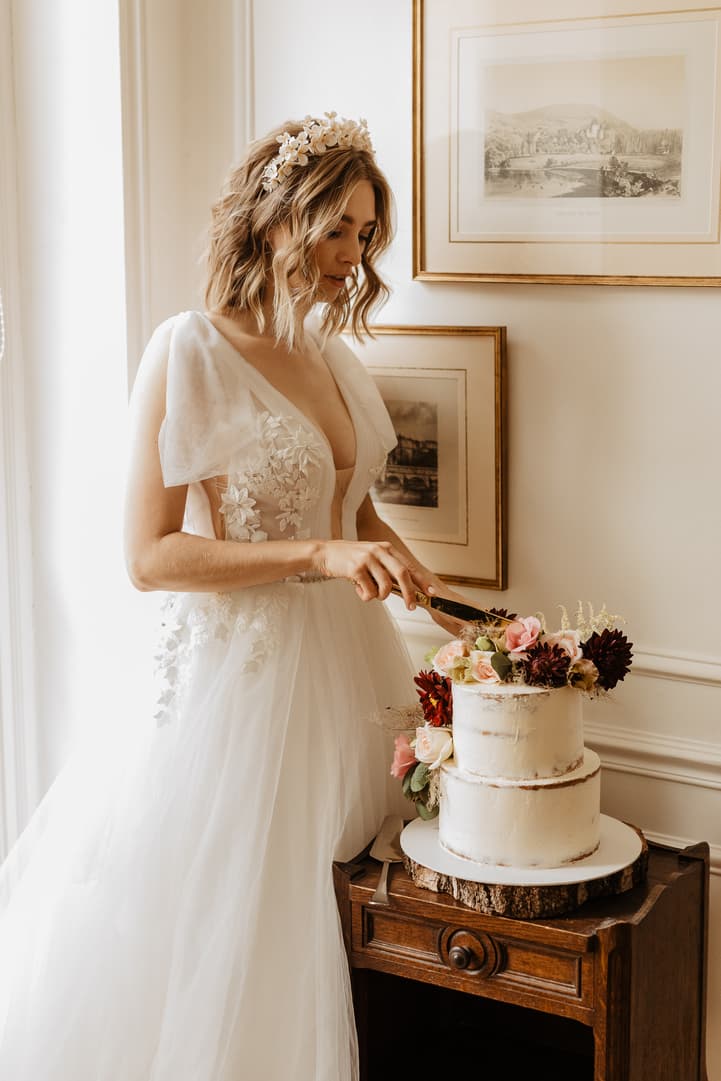 Bride in a hotel room cuts a two-tier cake topped with flowers beside soft window light