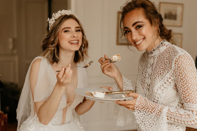 Two women in white lace dresses smile and share cake in a softly lit hotel room
