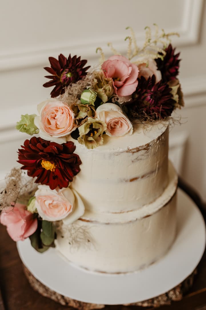 Two-tier wedding cake topped with blush and burgundy flowers on a table in soft indoor light