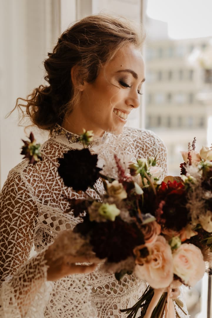 Bride in lace dress smiles by a bright window, holding a lush bouquet in a Paris hotel room