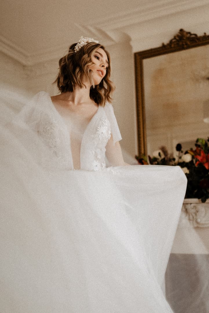 Bride in a flowing white gown stands in soft window light inside a Paris apartment