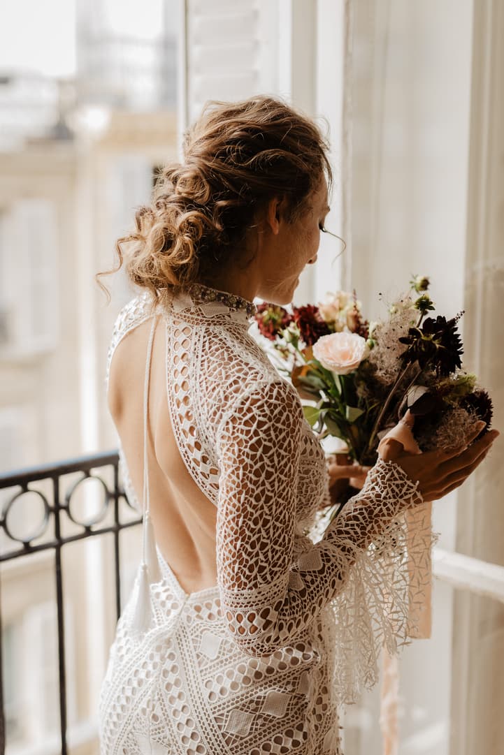 Bride in a lace backless gown holds a bouquet by a bright window in a Paris hotel room