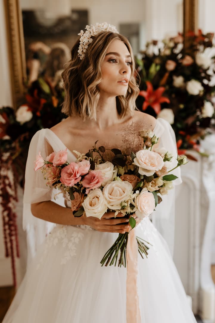 Bride in a white gown holds a pastel bouquet in soft indoor light, floral backdrop behind her