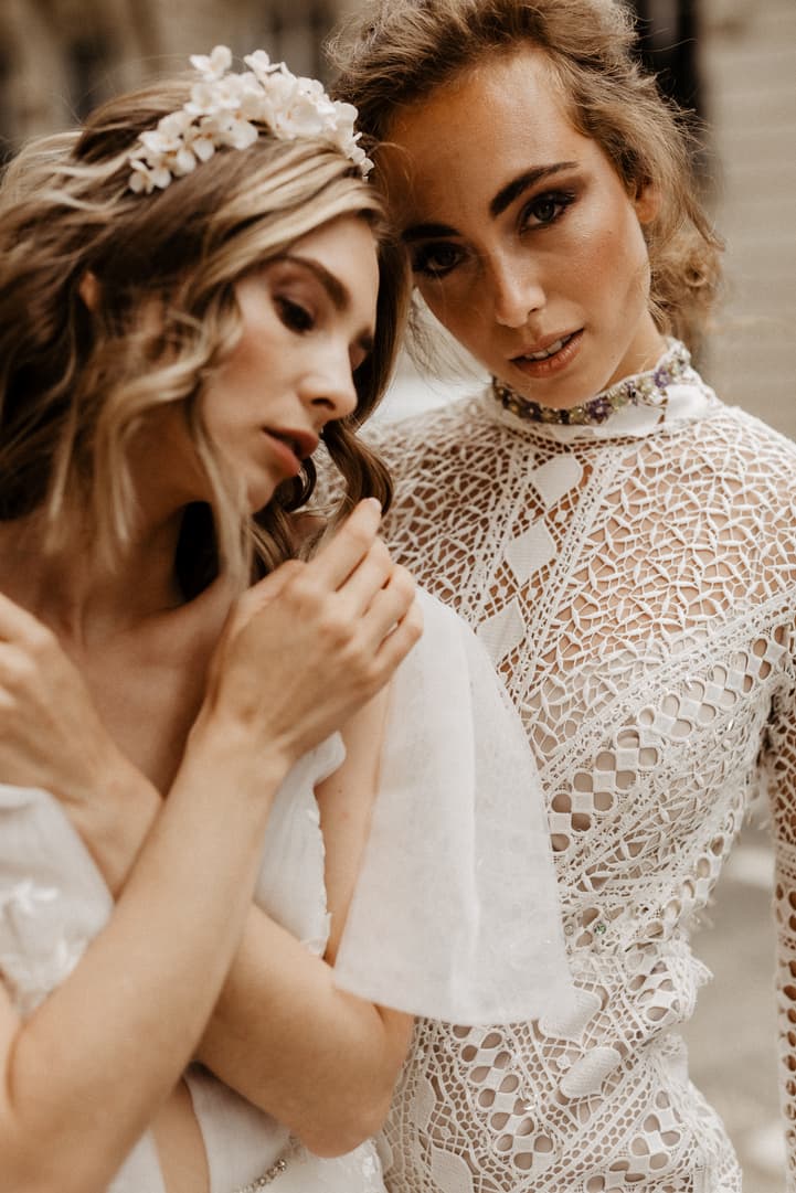 Two women in bridal lace pose closely in soft indoor light, one adjusting her hair