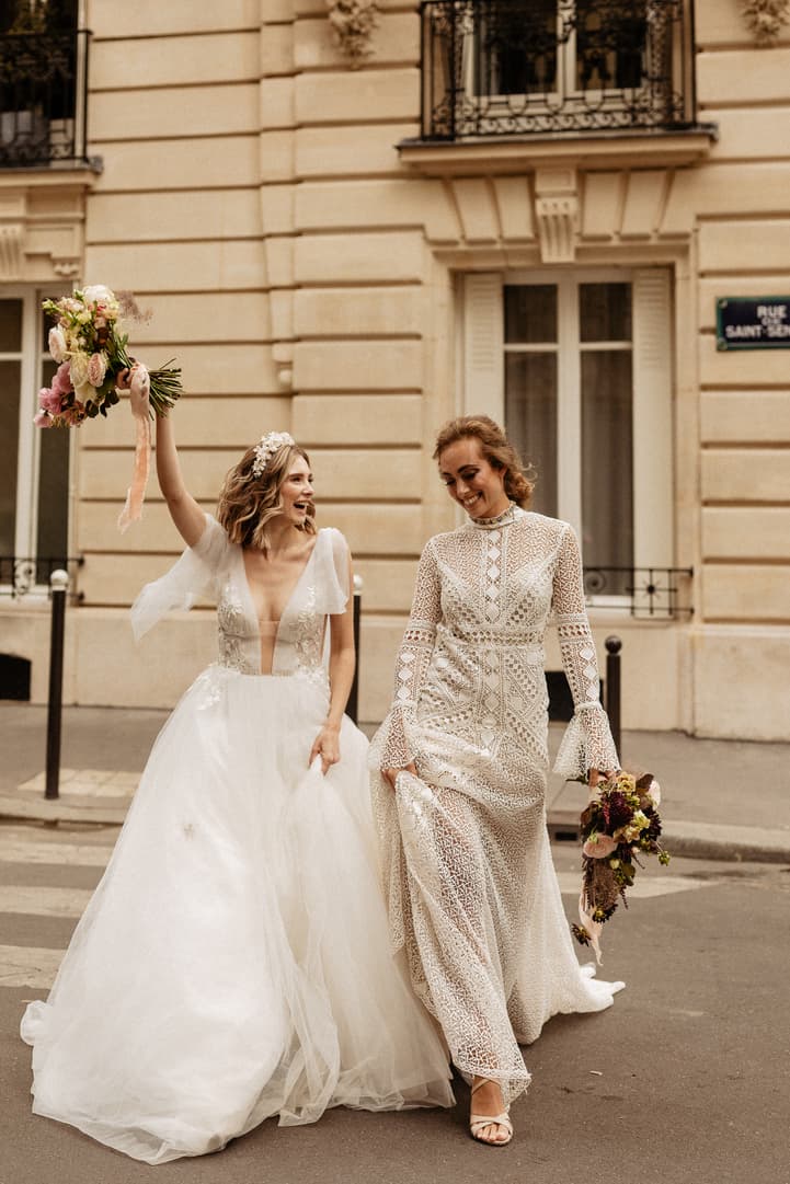 Two brides walk hand in hand on a Paris street, bouquets raised in warm light