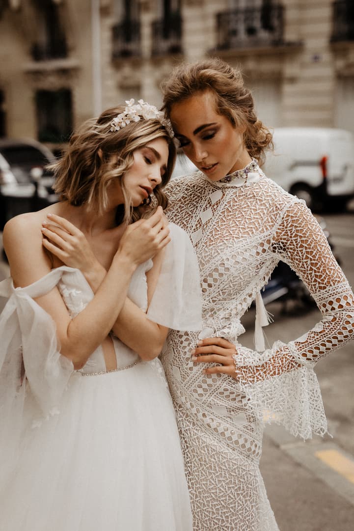 Two brides in lace gowns lean together on a Paris street in soft light, one wearing a floral headpiece