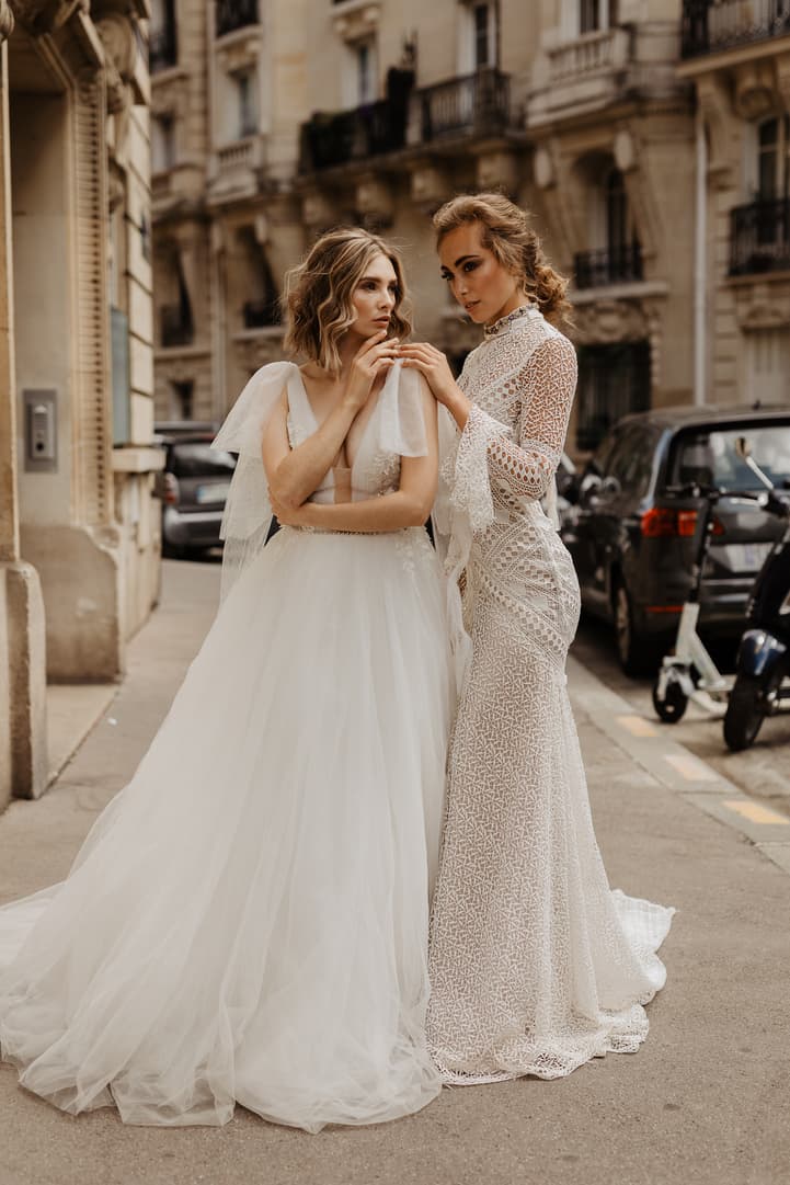 Bride in a flowing gown and lace-clad companion pose on a Paris street, hands on her shoulders in soft daylight