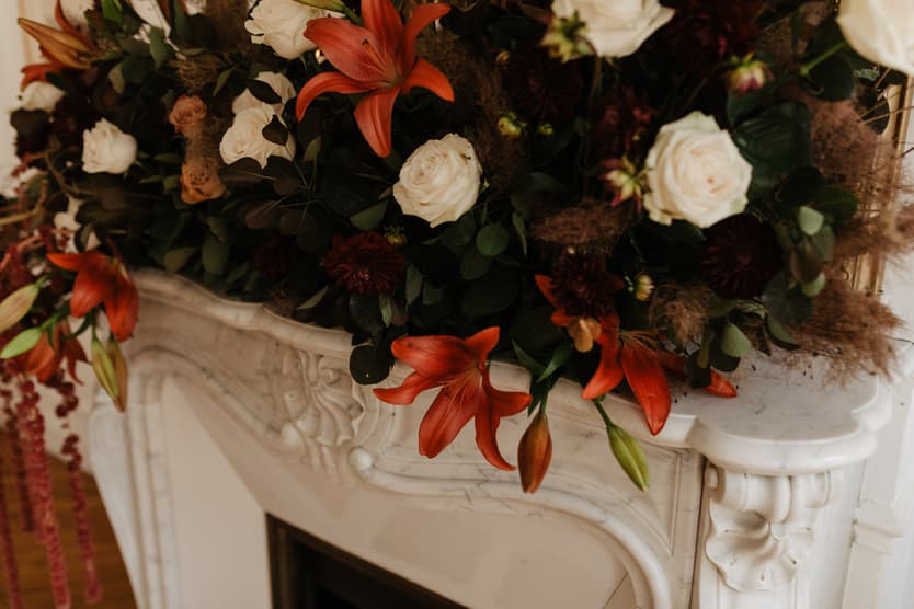 Orange lilies and white roses spill across a carved white mantel in soft indoor light