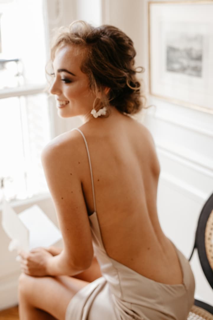 Bride in a backless satin dress smiles by a bright window in a Paris hotel room