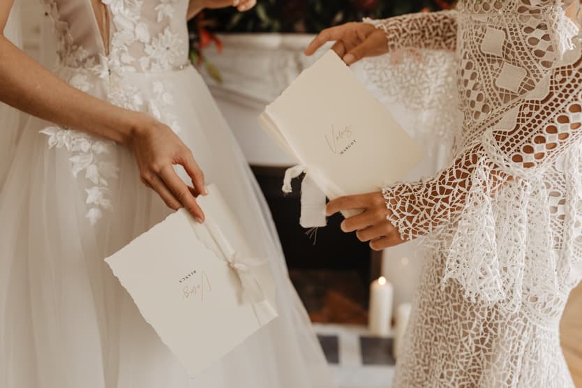 Bride and officiant in lace sleeves hold ceremony booklets by a candlelit fireplace