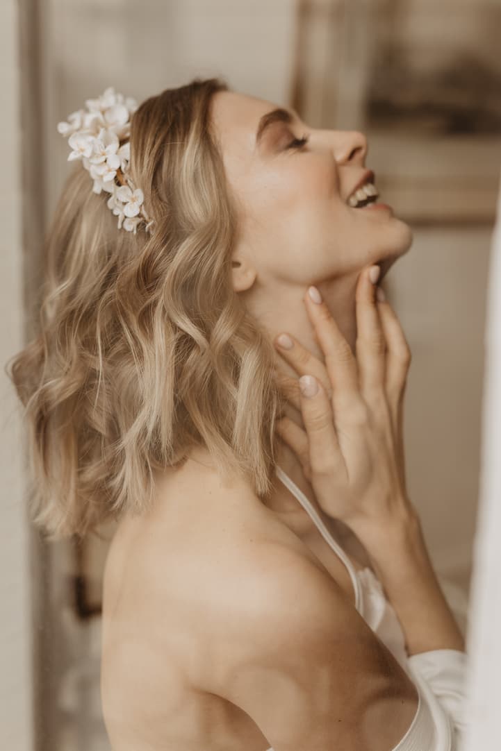 Bride in soft indoor light, smiling in profile with a floral hairpiece and hand at her neck