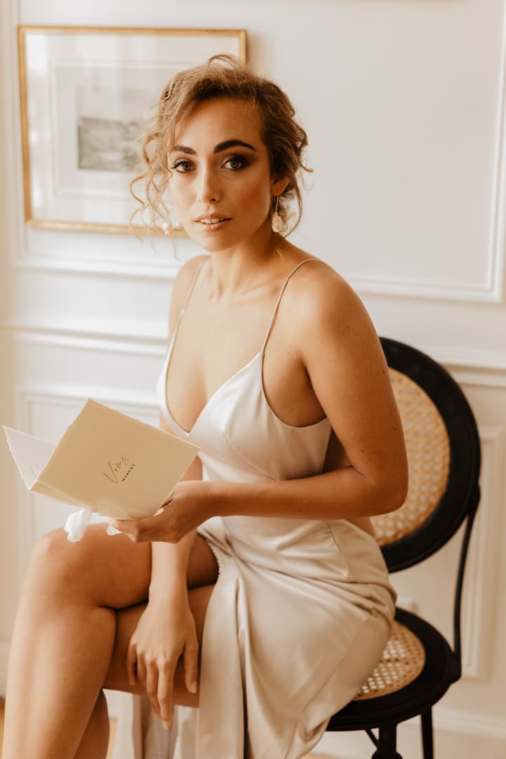 Bride in a satin slip sits by a cane chair, holding a card in warm window light