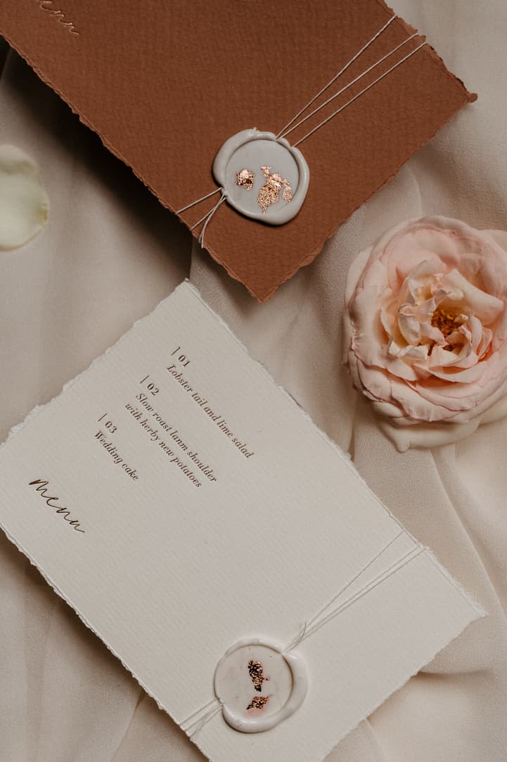Wedding menu and wax-sealed card on blush silk beside a pale pink rose in soft light