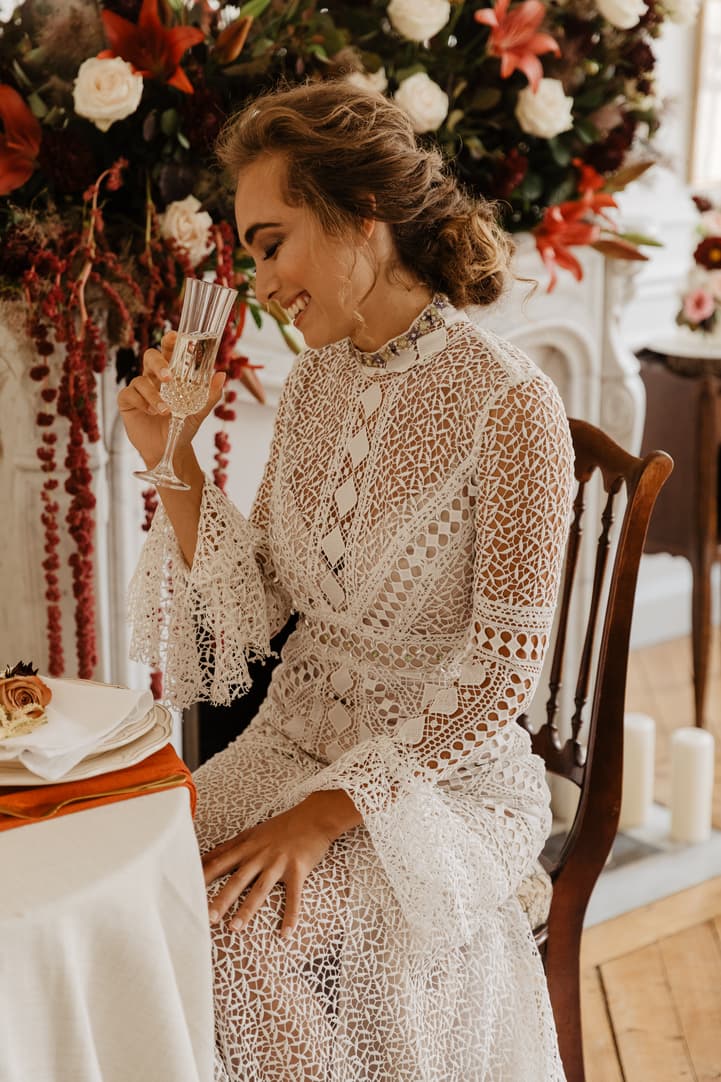 Bride in a lace dress raises a champagne flute at a floral table in warm window light