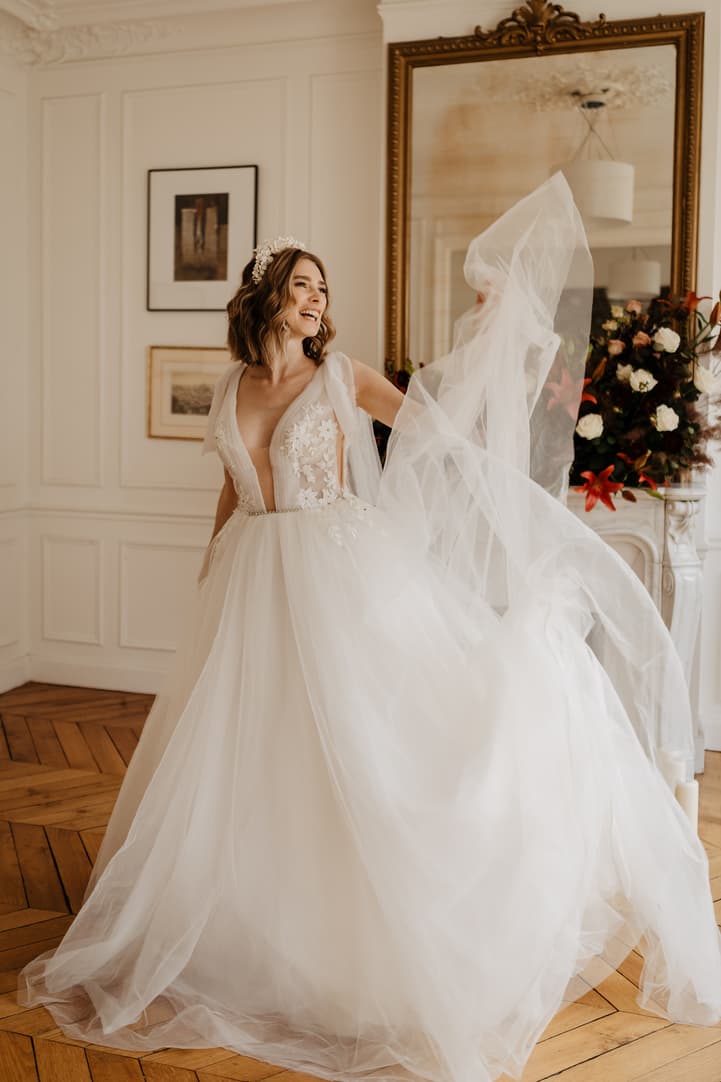 Bride in a flowing white gown lifts her veil in a sunlit Paris room with flowers and gilded mirror