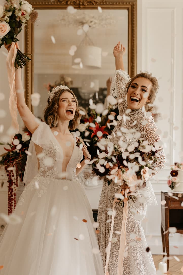 Two brides in lace dresses celebrate indoors as confetti falls and one holds a bouquet aloft