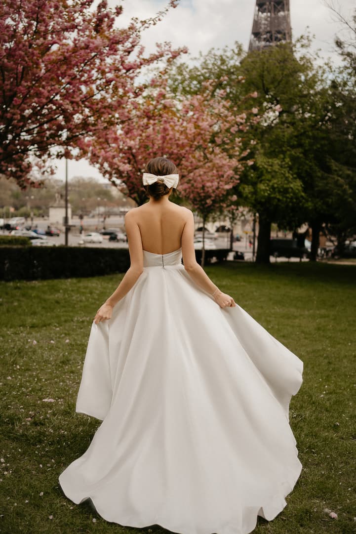 Bride in a white gown with a bow walks through a Paris garden under soft light, Eiffel Tower behind