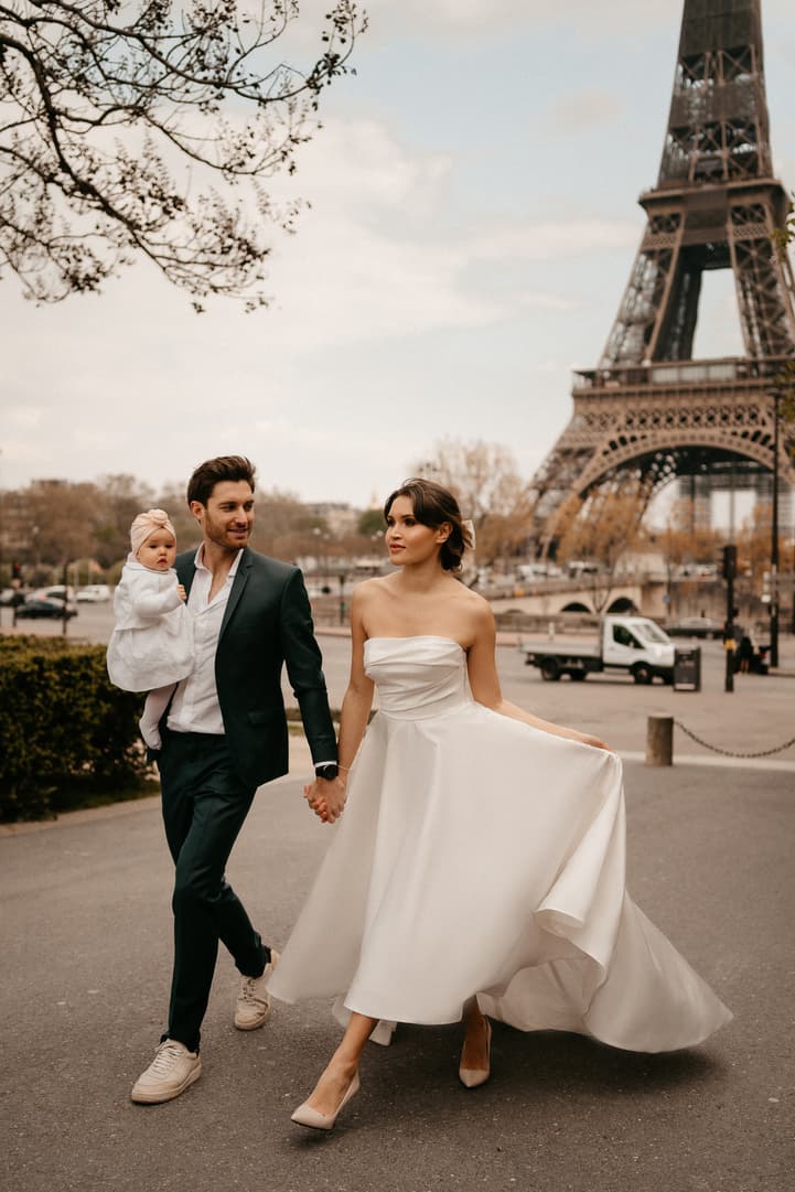 Bride and groom walk by the Eiffel Tower, with a child in his arms and her dress flowing