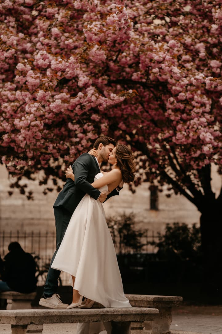 Bride and groom kiss on a stone bench beneath pink blossoms in a Paris garden