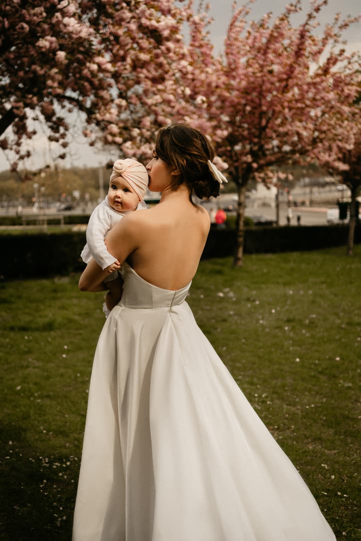 Bride in a white gown holds a baby in a blossom-filled garden near a Paris street