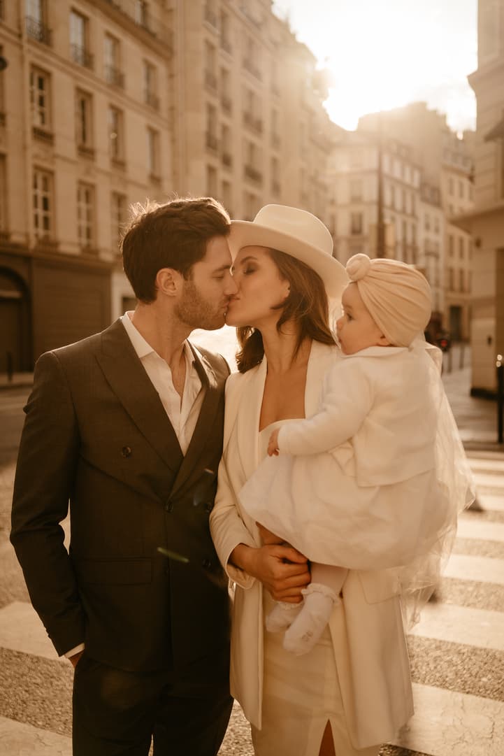 Bride and groom kiss while holding a baby on a sunlit Paris street, backlit by warm late-day light
