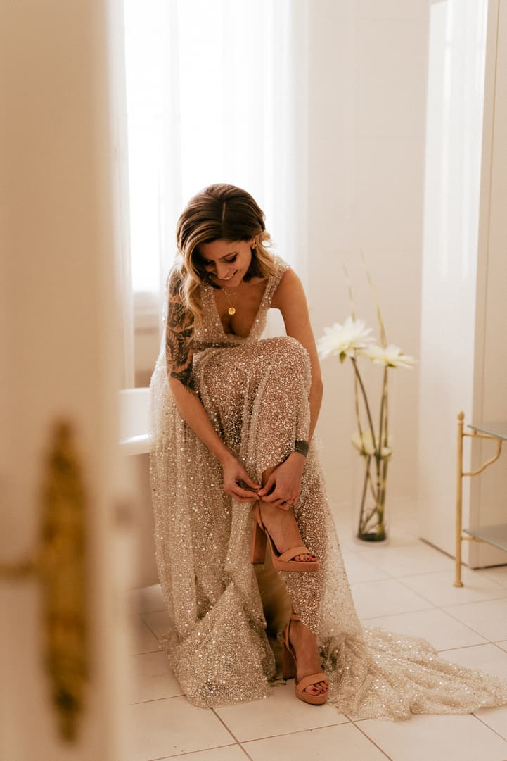 Bride in a sparkling gown adjusts her shoe in a softly lit hotel room beside white flowers