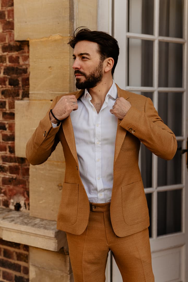 Groom adjusts his camel suit beside a Paris window in soft light