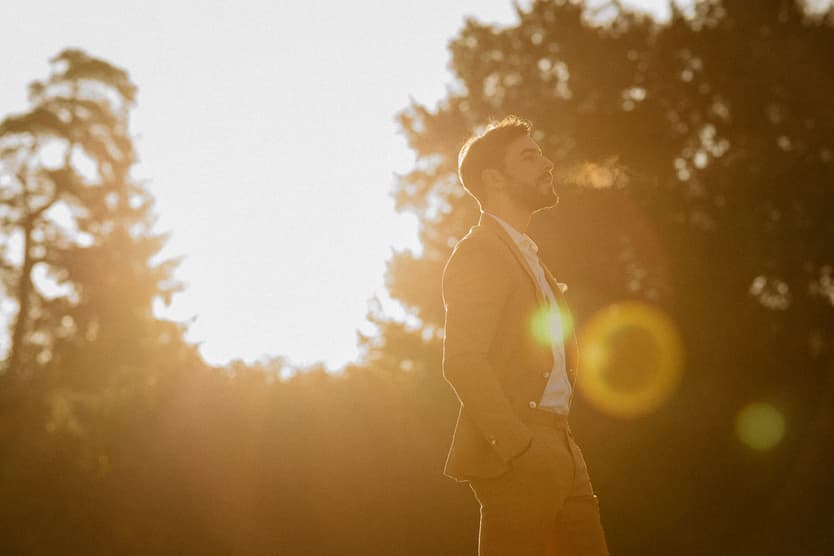 Groom in a suit stands in warm sunset light in a Paris garden, with lens flare and trees behind him