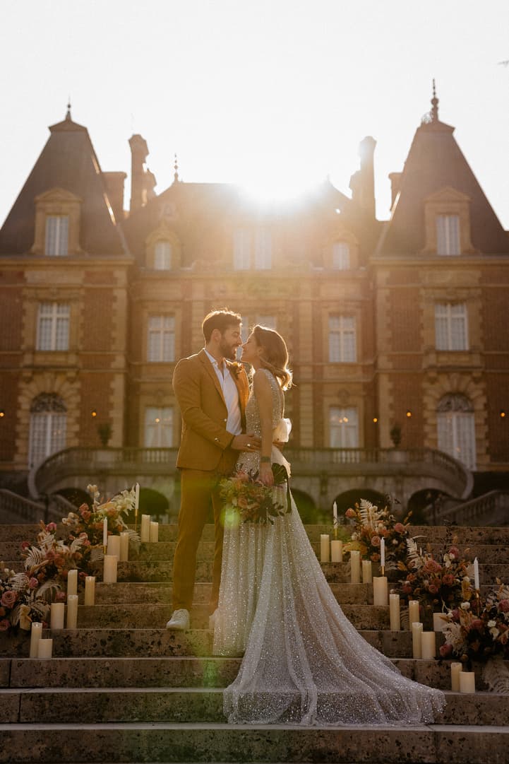 Bride and groom stand on flower-lined steps before a Paris chateau in warm sunset light and candles
