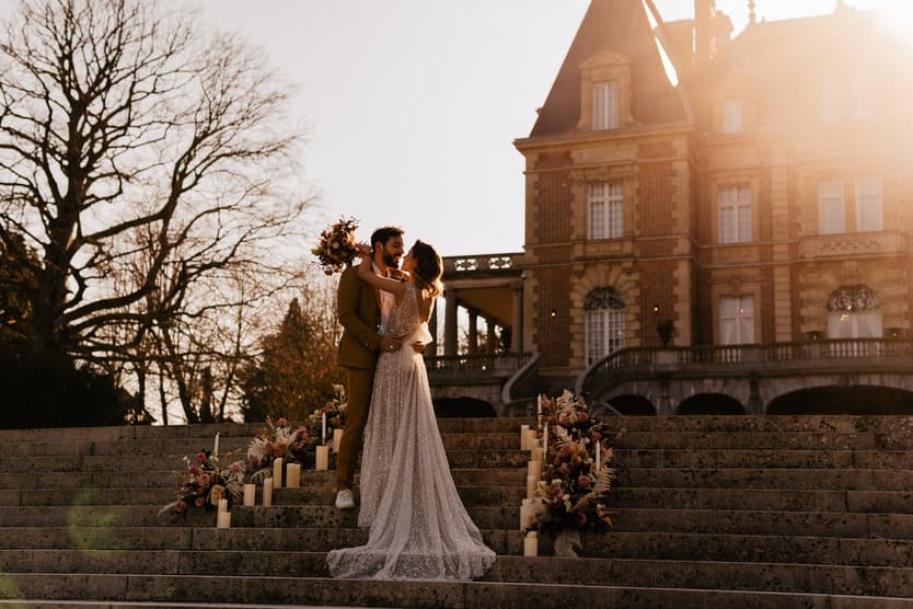 Bride and groom embrace on flower-lined stone steps outside a Paris mansion in warm sunset light