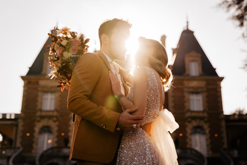 Bride and groom kiss in warm backlight before a Paris château, bouquet over his shoulder