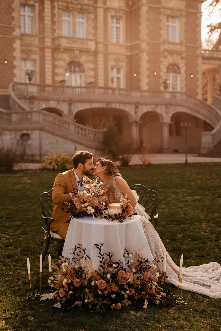Bride and groom kiss at a candlelit table in a garden, with floral decor and warm evening light before a grand Paris building